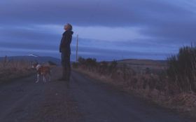 A woman stands on a country road at twilight, looking off into the distance, with a dog at her feet and snow-topped hills in the distance.